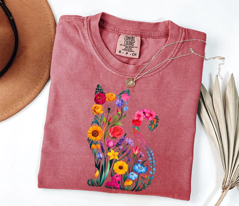 Op de afbeelding: Een stoffig roze Comfort Colors t-shirt met een bloemenkatontwerp. De kat is samengesteld uit kleurrijke bloemen, waaronder zonnebloemen, rozen en blauwe klokjes. Een zilveren ketting met een hanger rust op het shirt. Het shirt is maat S-P-CH.