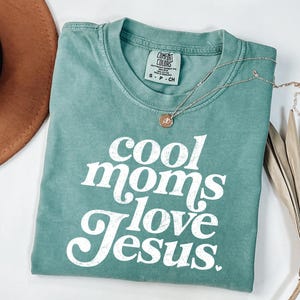 May include: A teal Comfort Colors t-shirt with the text "cool moms love Jesus" in white script. A gold necklace with a small pendant rests on the shirt. A brown hat and dried palm leaves are in the background.