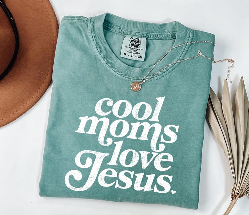 May include: A teal Comfort Colors t-shirt with the text "cool moms love Jesus" in white script. A gold necklace with a small pendant rests on the shirt. A brown hat and dried palm leaves are in the background.