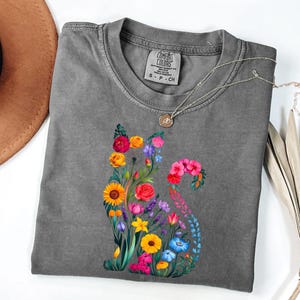 Op de afbeelding: Een grijs Comfort Colors T-shirt met een bloemenkatontwerp. De kat is samengesteld uit kleurrijke bloemen, waaronder zonnebloemen, rozen en blauwe klokjes. Een zilveren ketting met een hanger ligt op het shirt.