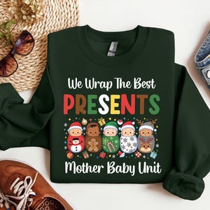 May include: Dark green sweatshirt with the text "We Wrap The Best PRESENTS Mother Baby Unit." The design features illustrations of babies dressed as Christmas characters, including a snowman, gingerbread man, and reindeer.