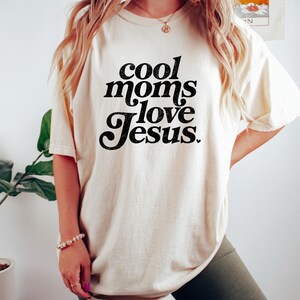 May include: Cream-colored t-shirt with the text "cool moms love Jesus" in black script. The shirt is a casual style, suitable for everyday wear. The image shows the shirt being worn.