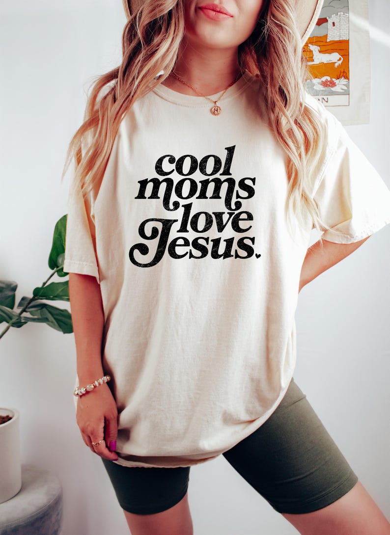 May include: Cream-colored t-shirt with the text "cool moms love Jesus" in black script. The shirt is a casual style, suitable for everyday wear. The image shows the shirt being worn.