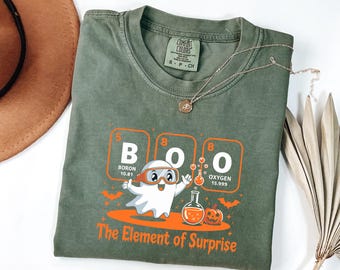 Può includere: T-shirt verde oliva con una grafica raffigurante un fantasma con occhiali e la parola "BOO" in riquadri della tavola periodica. Il testo "The Element of Surprise" è sotto la grafica. Una collana d'oro e un cappello marrone sono sullo sfondo.