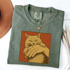 以下が含まれることがあります： 猫が手を噛んでいるグラフィックが特徴のセージグリーンのTシャツ。猫は黄色で、手はタン色で、赤い背景に設定されています。小さなペンダントが付いたシルバーのネックレスがシャツの上にかけられています。