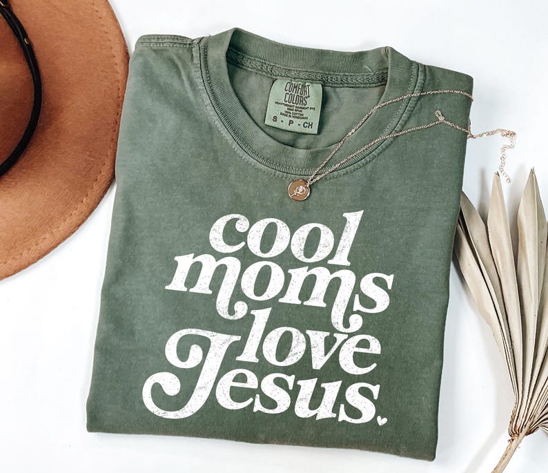 May include: An olive green t-shirt with the text "cool moms love Jesus" in white, a gold necklace, and a brown hat. The shirt is made of a soft material and has a crew neck.