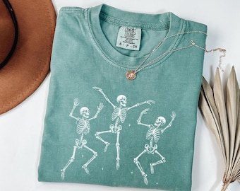 May include: A teal Comfort Colors t-shirt featuring a white graphic of three dancing skeletons. A gold necklace with a pendant rests on the shirt. The shirt is folded, with a brown hat and dried plant in the background.