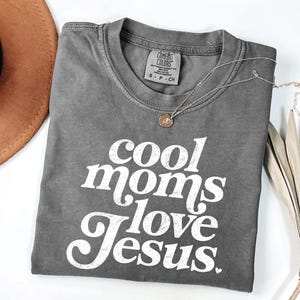 May include: A gray Comfort Colors t-shirt with the text "cool moms love Jesus" in white. A gold necklace with a pendant rests on the shirt. A brown hat and dried plant are in the background.
