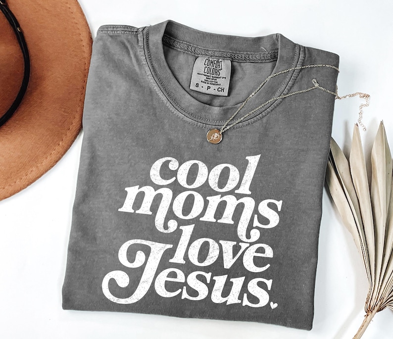 May include: A gray Comfort Colors t-shirt with the text "cool moms love Jesus" in white. A gold necklace with a pendant rests on the shirt. A brown hat and dried plant are in the background.