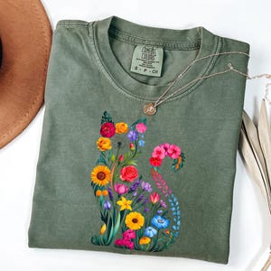 Op de afbeelding: Een olijfgroen t-shirt met een bloemenkatontwerp. De kat is samengesteld uit kleurrijke bloemen, waaronder zonnebloemen, rozen en klokjes. Een zilveren ketting met een kleine hanger rust op het shirt. Het shirt is van het merk Comfort Colors.