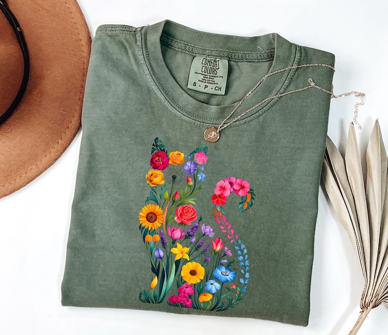 Op de afbeelding: Een olijfgroen t-shirt met een bloemenkatontwerp. De kat is samengesteld uit kleurrijke bloemen, waaronder zonnebloemen, rozen en klokjes. Een zilveren ketting met een kleine hanger rust op het shirt. Het shirt is van het merk Comfort Colors.