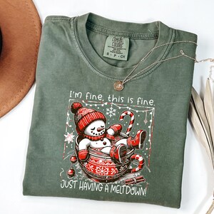以下が含まれることがあります： オリーブグリーンのTシャツ。カップに入った雪だるまのグラフィックで、赤い帽子とミトンを着用しています。「I'm fine, this is fine. JUST HAVING A MELT DOWN!」と書かれています。金のネックレスがシャツの上にかけられています。