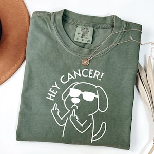 May include: An olive green t-shirt with the text "HEY CANCER!" and a cartoon dog wearing sunglasses and giving the middle finger. A gold necklace with a pendant is draped across the shirt.