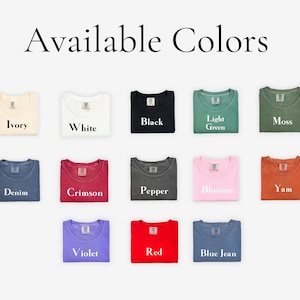 Op de afbeelding: Een display van opgevouwen t-shirts in verschillende kleuren, waaronder ivoor, wit, zwart, lichtgroen en mos. Andere kleuren zijn denim, karmozijnrood, peper, bloesem, yam, violet, rood en blue jean. De tekst "Beschikbare kleuren" staat bovenaan.