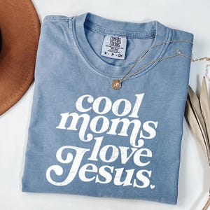 May include: A blue Comfort Colors t-shirt with the text "cool moms love Jesus" in white. A gold necklace with a pendant is draped across the shirt. A brown hat is visible in the upper left corner.