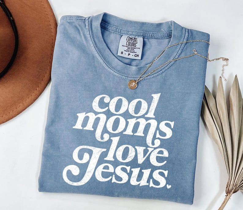 May include: A blue Comfort Colors t-shirt with the text "cool moms love Jesus" in white. A gold necklace with a pendant is draped across the shirt. A brown hat is visible in the upper left corner.