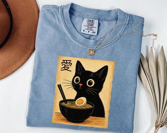May include: A light blue Comfort Colors t-shirt featuring a graphic of a black cat eating ramen. The graphic includes the Japanese character for love. A gold necklace is draped over the shirt.