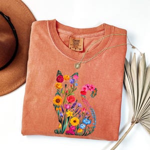 Op de afbeelding: Een koraalkleurig Comfort Colors T-shirt met een bloemenkatontwerp. De kat bestaat uit kleurrijke bloemen, waaronder zonnebloemen, rozen en lavendel. Een gouden ketting en een bruine hoed staan ook op de foto.