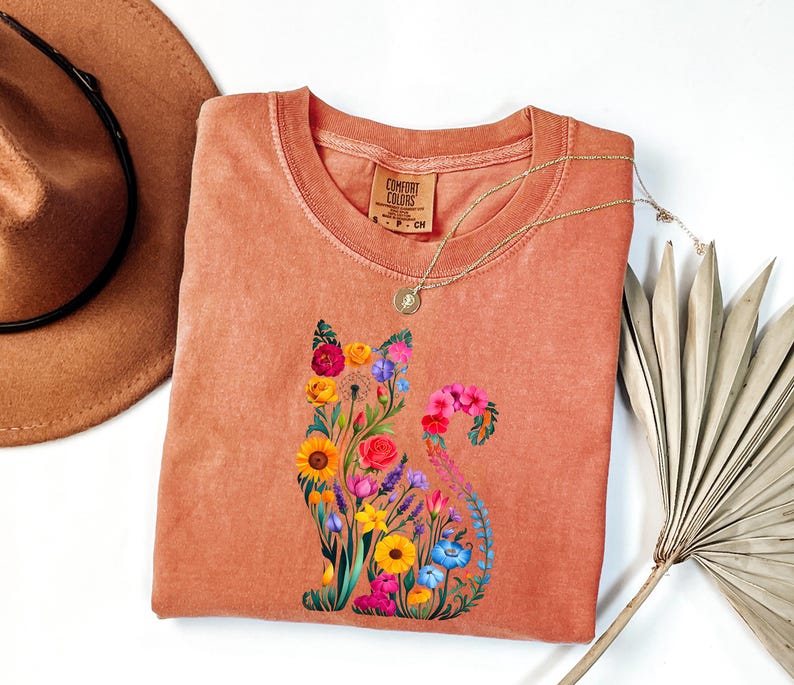 Op de afbeelding: Een koraalkleurig Comfort Colors T-shirt met een bloemenkatontwerp. De kat bestaat uit kleurrijke bloemen, waaronder zonnebloemen, rozen en lavendel. Een gouden ketting en een bruine hoed staan ook op de foto.