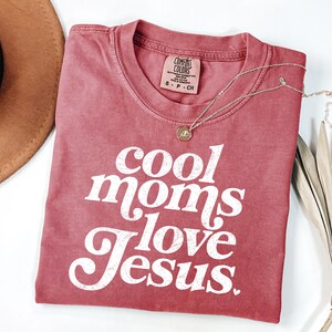 May include: A red Comfort Colors t-shirt with the text "cool moms love Jesus" in white script. A gold necklace with a pendant is draped across the shirt. A brown hat and dried plant are in the background.