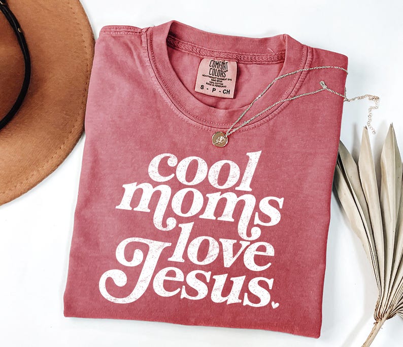 May include: A red Comfort Colors t-shirt with the text "cool moms love Jesus" in white script. A gold necklace with a pendant is draped across the shirt. A brown hat and dried plant are in the background.