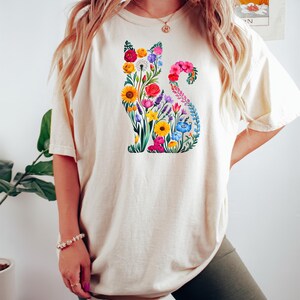 Op de afbeelding: Cr&egrave;mekleurig T-shirt met een bloemenkatontwerp. Het katten silhouet is gevuld met kleurrijke bloemen, waaronder gele zonnebloemen, rode rozen en blauwe bloemen. Het shirt heeft korte mouwen en een relaxte pasvorm.