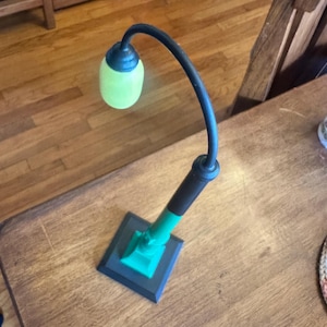 Puede incluir: Una pequeña lámpara decorativa con un brazo negro curvado y una pantalla verde redondeada. La base tiene forma cuadrada con una combinación de colores verde y negro. La lámpara está sobre una superficie de madera.