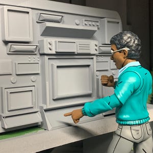 Op de afbeelding: Een grijs bedieningspaneel met diverse rechthoekige en vierkante componenten. Een figuur in een turquoise trui en een bril wijst naar het paneel. Het paneel heeft een scherm en diverse knoppen.