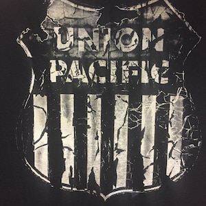 Pode incluir: Camiseta preta com um gráfico branco desgastado do logotipo da Union Pacific. O logotipo em forma de escudo apresenta listras verticais e as palavras "UNION PACIFIC" em um estilo vintage e desgastado. O design tem uma aparência rachada e envelhecida.