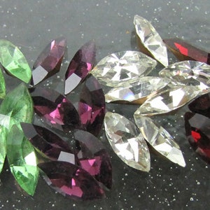 Puede incluir: Un surtido de gemas de talla marquesa en un arcoíris de colores, incluyendo verde, amarillo, morado, transparente, rojo y azul. Las gemas están dispuestas en un patrón decorativo, mostrando sus facetas y brillo.