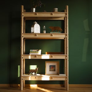 May include: A natural wood bookshelf with several shelves, showcasing decorative items such as a lantern, figurines, books, and framed art. The shelves are filled with various objects, creating a visually appealing display against a dark green wall.