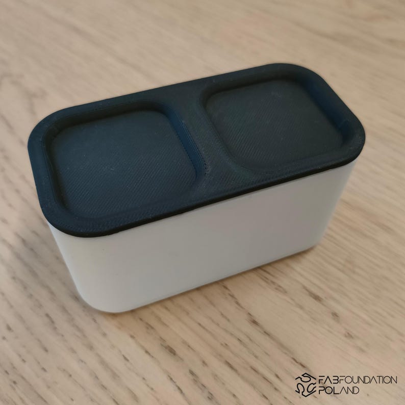 Pu&ograve; includere: Un contenitore rettangolare bianco con un coperchio nero a due scomparti. Il coperchio ha una superficie strutturata. Il contenitore &egrave; su una superficie di legno chiaro.