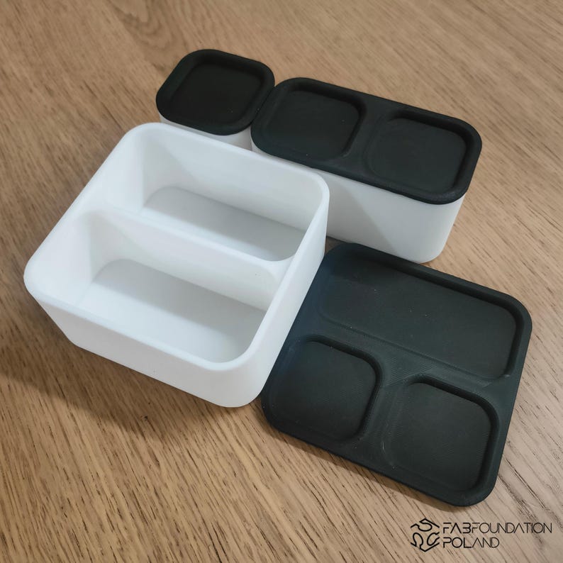 Pu&ograve; includere: Contenitori per alimenti in plastica bianca e nera con coperchi. Un contenitore &egrave; diviso in due scomparti. I coperchi sono neri e hanno forma rettangolare o quadrata. I contenitori sono su una superficie di legno.