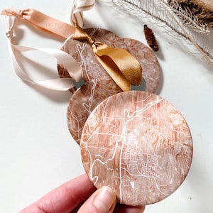 May include: Two round wooden ornaments with a white map design. The ornaments are tied with a brown and white striped ribbon, a light pink ribbon, and a gold ribbon.