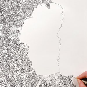 Op de afbeelding: Een gedetailleerde zwart-wit topografische kaarttekening op wit papier. De kaart toont gedetailleerde landformaties en een kustlijn met ingewikkelde lijnen. Een hand met een potlood is zichtbaar, wat het artistieke proces aangeeft.