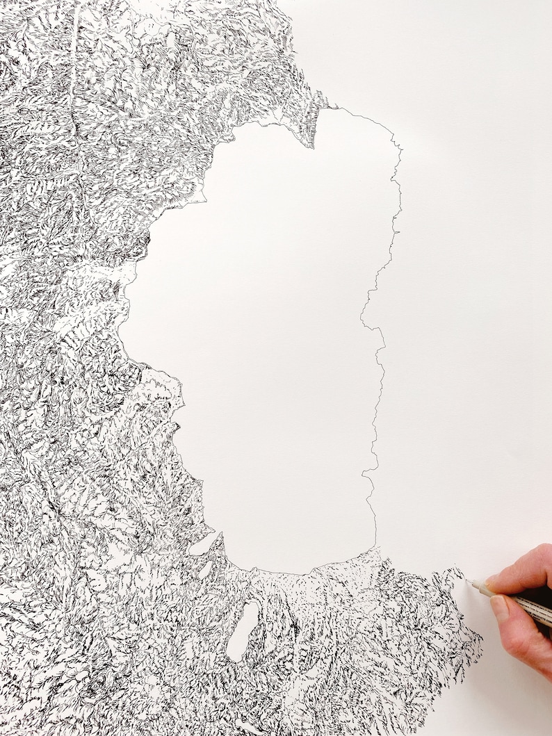 Op de afbeelding: Een gedetailleerde zwart-wit topografische kaarttekening op wit papier. De kaart toont gedetailleerde landformaties en een kustlijn met ingewikkelde lijnen. Een hand met een potlood is zichtbaar, wat het artistieke proces aangeeft.