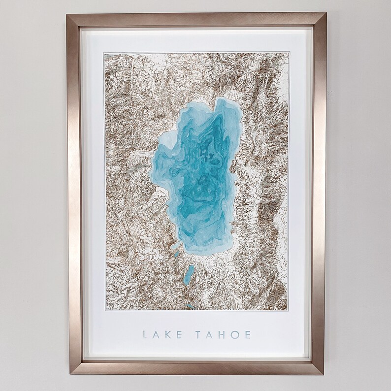 Op de afbeelding: Ingelijste kunstdruk van Lake Tahoe. Het kunstwerk toont een gedetailleerde topografische kaart van het meer in tinten blauw en bruin, met de naam van het meer in het wit onderaan. De print is ingekaderd in een metalen frame.