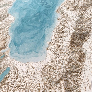 Op de afbeelding: Een gedetailleerde topografische kaart met een lichtblauw aquarelmeer en omliggend land in goud en wit. De kaart heeft een driedimensionaal effect, met schaduwen die diepte aan het landschap toevoegen.