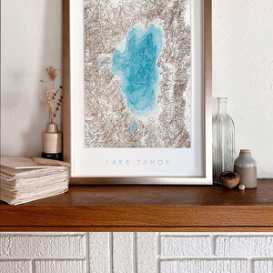 Op de afbeelding: Een ingelijste print van Lake Tahoe. Het kunstwerk toont een gedetailleerde topografische kaart van het meer in blauwe en bruine tinten. De print is geplaatst in een metalen frame en wordt getoond op een houten plank, met de tekst "LAKE TAHOE".