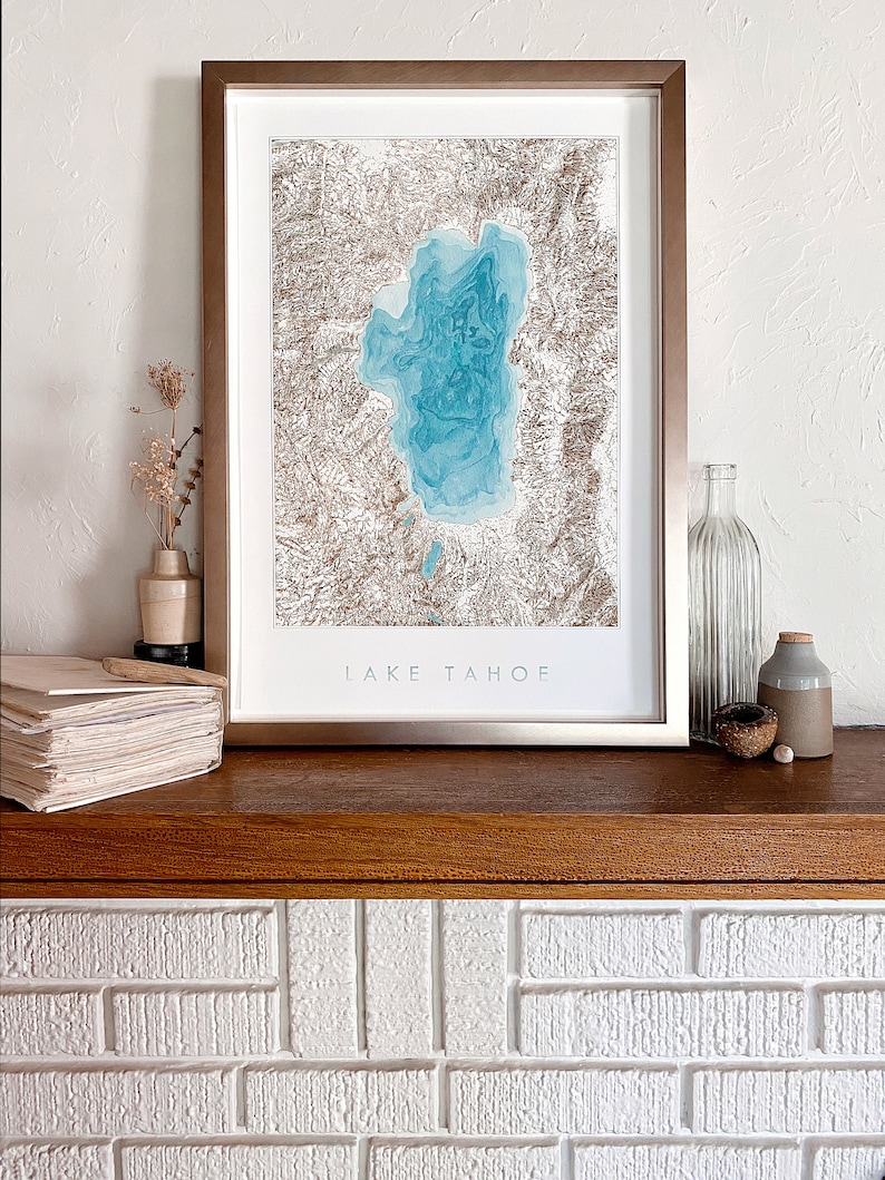 Op de afbeelding: Een ingelijste print van Lake Tahoe. Het kunstwerk toont een gedetailleerde topografische kaart van het meer in blauwe en bruine tinten. De print is geplaatst in een metalen frame en wordt getoond op een houten plank, met de tekst "LAKE TAHOE".
