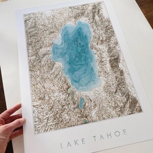 Op de afbeelding: Een print van Lake Tahoe, met een gedetailleerde topografische kaart in bruine en blauwe tinten. Het meer is weergegeven in turquoise, met de tekst "LAKE TAHOE" gedrukt in een lichtgrijze lettertype onderaan de witte rand.