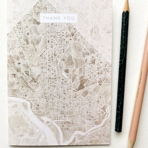 May include: A thank you card featuring a map design in shades of gray and white. The card has the words "THANK YOU" in a rectangular box. A black and a tan colored pencil are next to the card.
