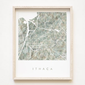 May include: A framed print of a watercolor map of Ithaca, New York. The map is in shades of green and brown, with white lines representing streets and roads. The word "ITHACA" is printed in white at the bottom of the print.