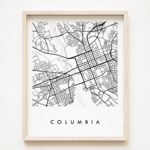 May include: A black and white line art map print of Columbia, South Carolina. The print is framed in a light wood frame and has the word "COLUMBIA" printed in black at the bottom.