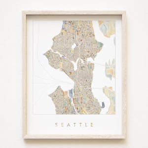 May include: A framed watercolour map of Seattle, Washington, showing a detailed depiction of the city's layout. The map is rendered in muted earth tones with gold lettering that spells "SEATTLE" at the bottom. The frame is a light wood.