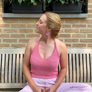 May include: A woman wearing a pink halter top and pink striped pants sits on a wooden bench. She is wearing pink rectangular earrings.