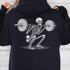 Könnte beinhalten: Schwarzer Hoodie mit einem weißen Grafikdruck eines Skeletts, das Gewichte hebt. Das Skelett trägt Turnschuhe und hebt eine Langhantel mit Gewichten an jedem Ende.