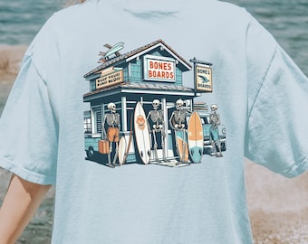 May include: A light blue t-shirt featuring a cartoon surf shop with skeletons holding surfboards. The shop is called "Bones Boards" and has a sign that says "Surf Tour Surf Board".