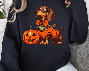 May include: A navy blue sweatshirt with a dachshund in a pumpkin costume and bow tie, next to a jack-o'-lantern. The dog has brown fur and wears an orange costume with bat wings. The jack-o'-lantern is orange with a carved face.