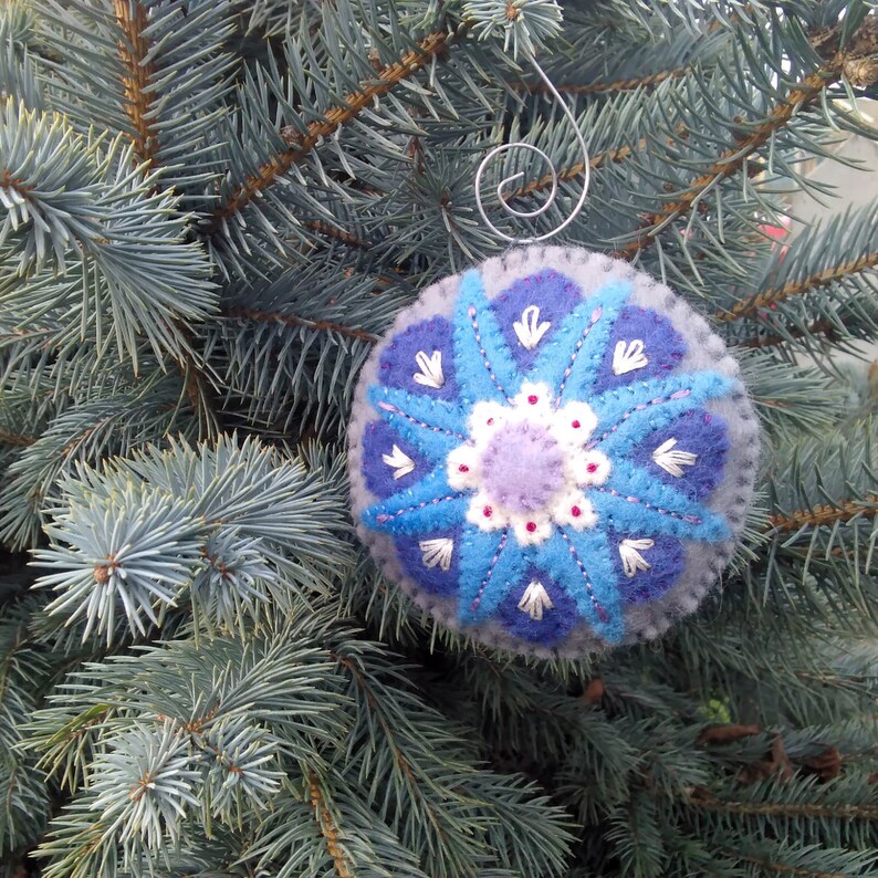 May include: A blue and purple felt ornament with a snowflake design. The ornament is hanging on a blue spruce tree.